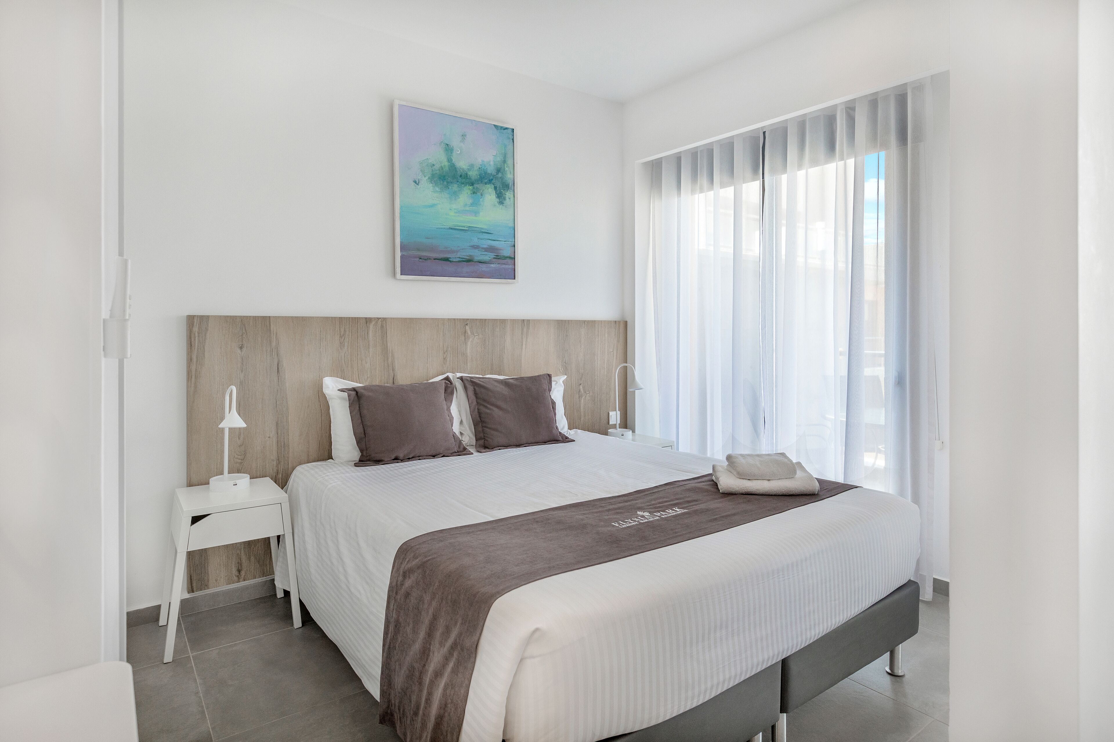Une chambre d’hôtel moderne avec un grand lit, une tête de lit en bois, une table de chevet avec une lampe, un tableau au mur et une fenêtre avec des rideaux légers.