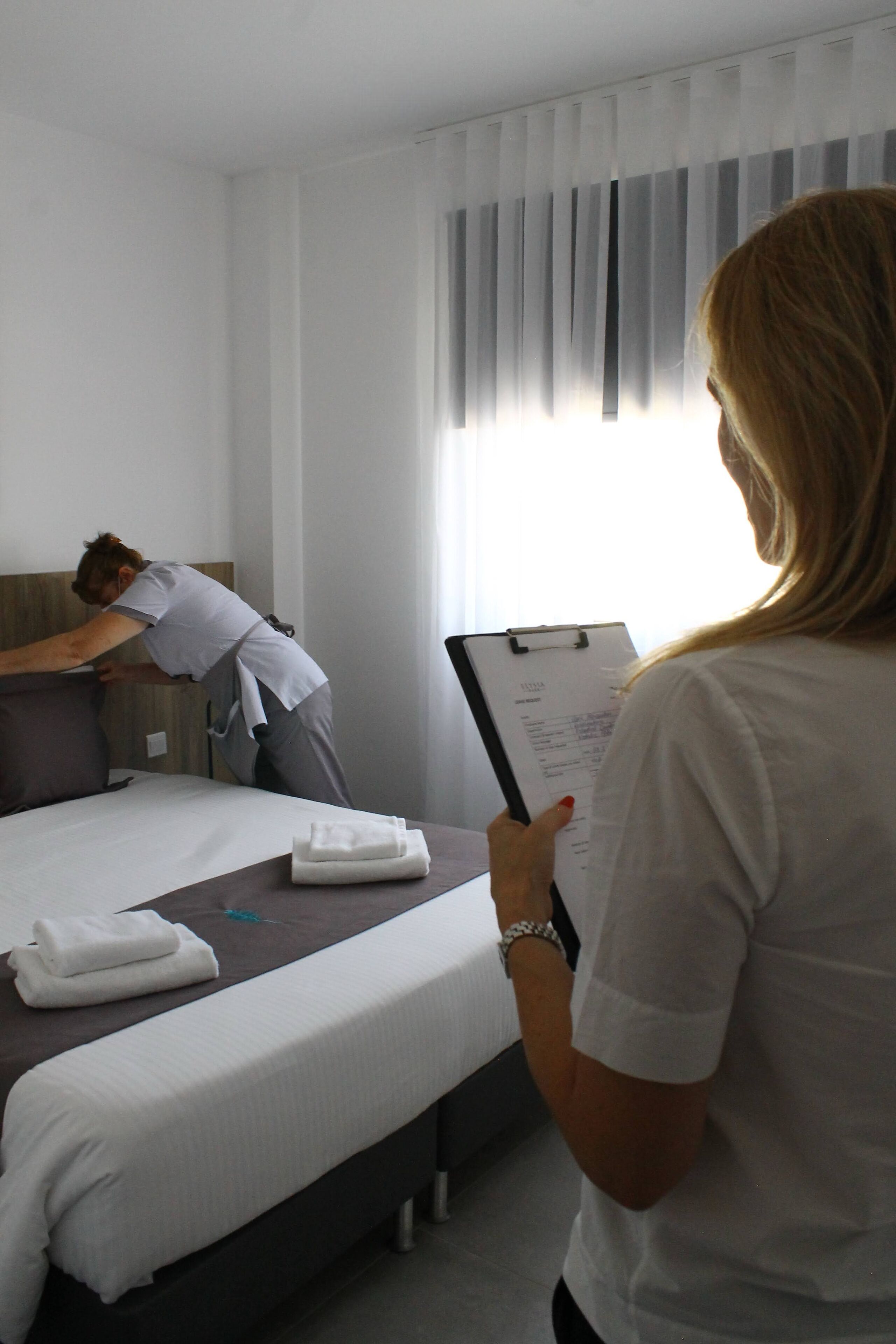 Een hotelkamer met een bed, nachtkastje, raam met zonwering, en een persoon die bij het bed staat.
