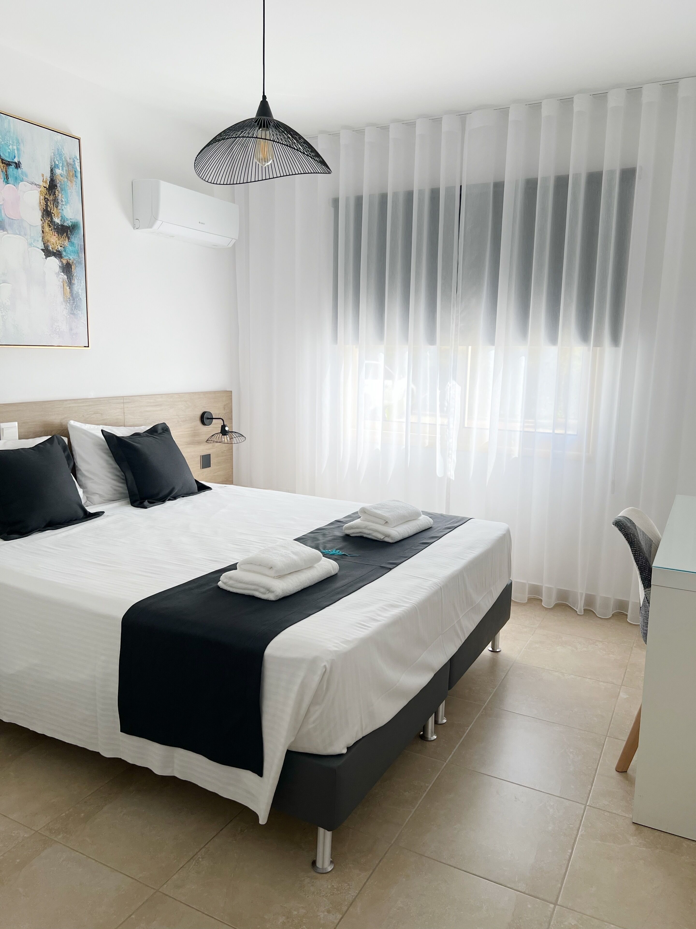 A modern hotel room with a large bed, white bedding, a black headboard, a bedside table with a lamp, and sheer curtains.
