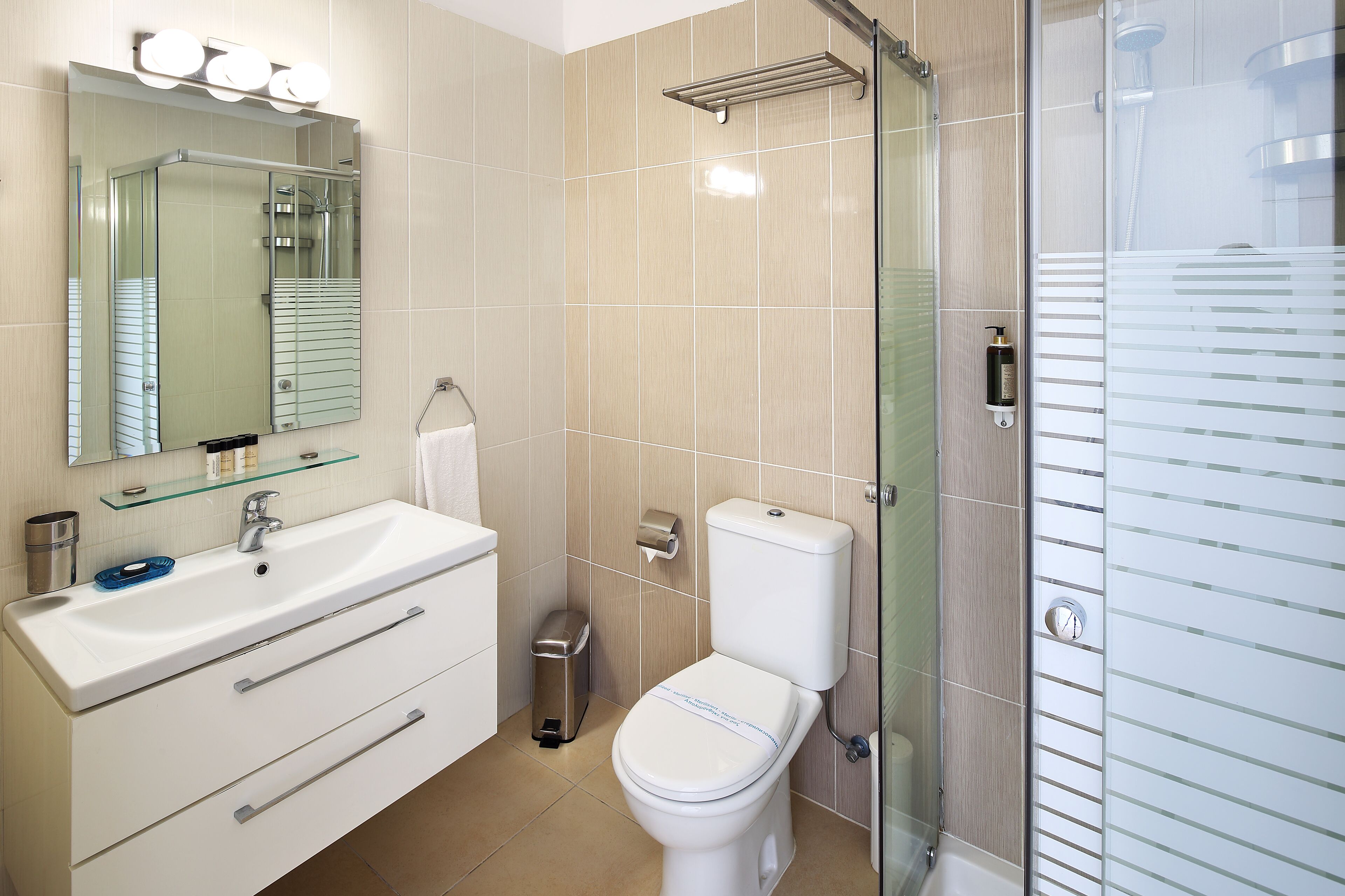 A modern bathroom with a glass shower enclosure, a white sink with a mirror above, and a toilet with a paper towel dispenser.