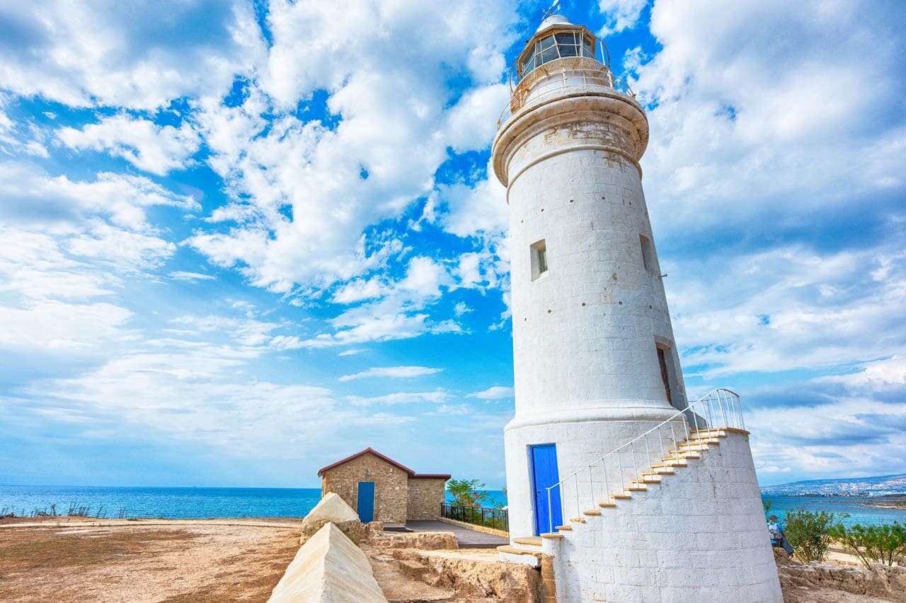 Un phare doté d’un escalier en colimaçon, un petit bâtiment adjacent avec une porte bleue, et une vue dégagée sur la mer sous un ciel partiellement nuageux.