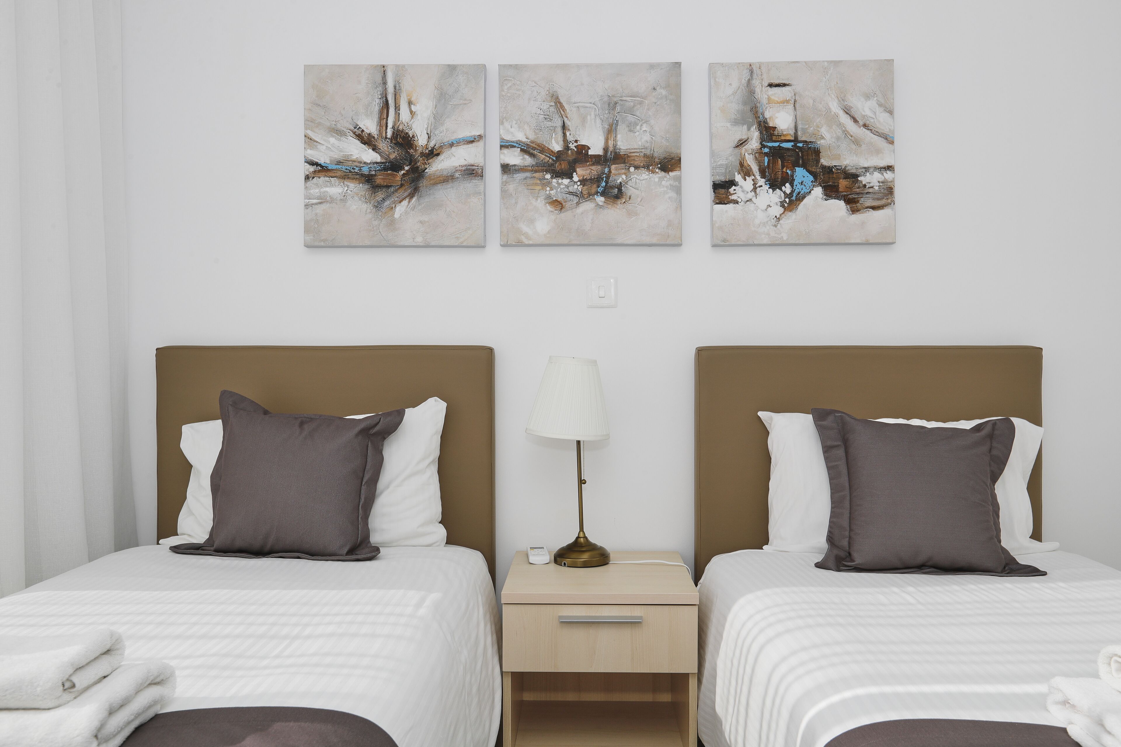A hotel room with two beds, a nightstand, and three abstract paintings on the wall.