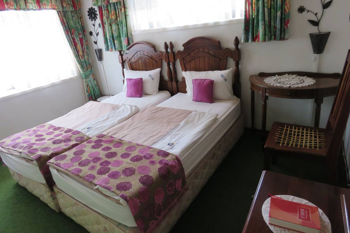 A double bed with two pillows, a small bedside table with a Bible, and a chair.
