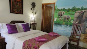 A bedroom with a bed, pillows, a nightstand, a lamp, a chair, and a mural of elephants. - Parrot Lodge (Bulawayo)