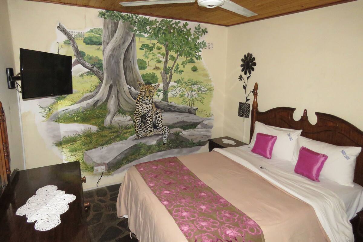 A bedroom with a bed, a nightstand, a TV, and a mural of a tree and leopard.