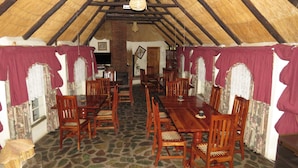 A dining area with wooden tables and chairs, a stone floor, and a thatched roof. - Parrot Lodge (Bulawayo)