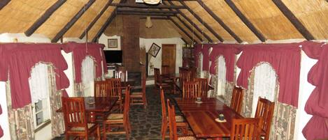 Un comedor con mesas y sillas de madera, suelo de piedra y techo de paja.