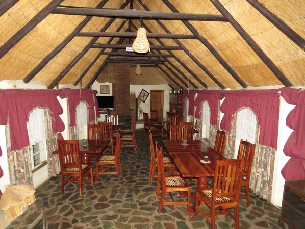Un comedor con mesas y sillas de madera, suelo de piedra y techo de paja.
