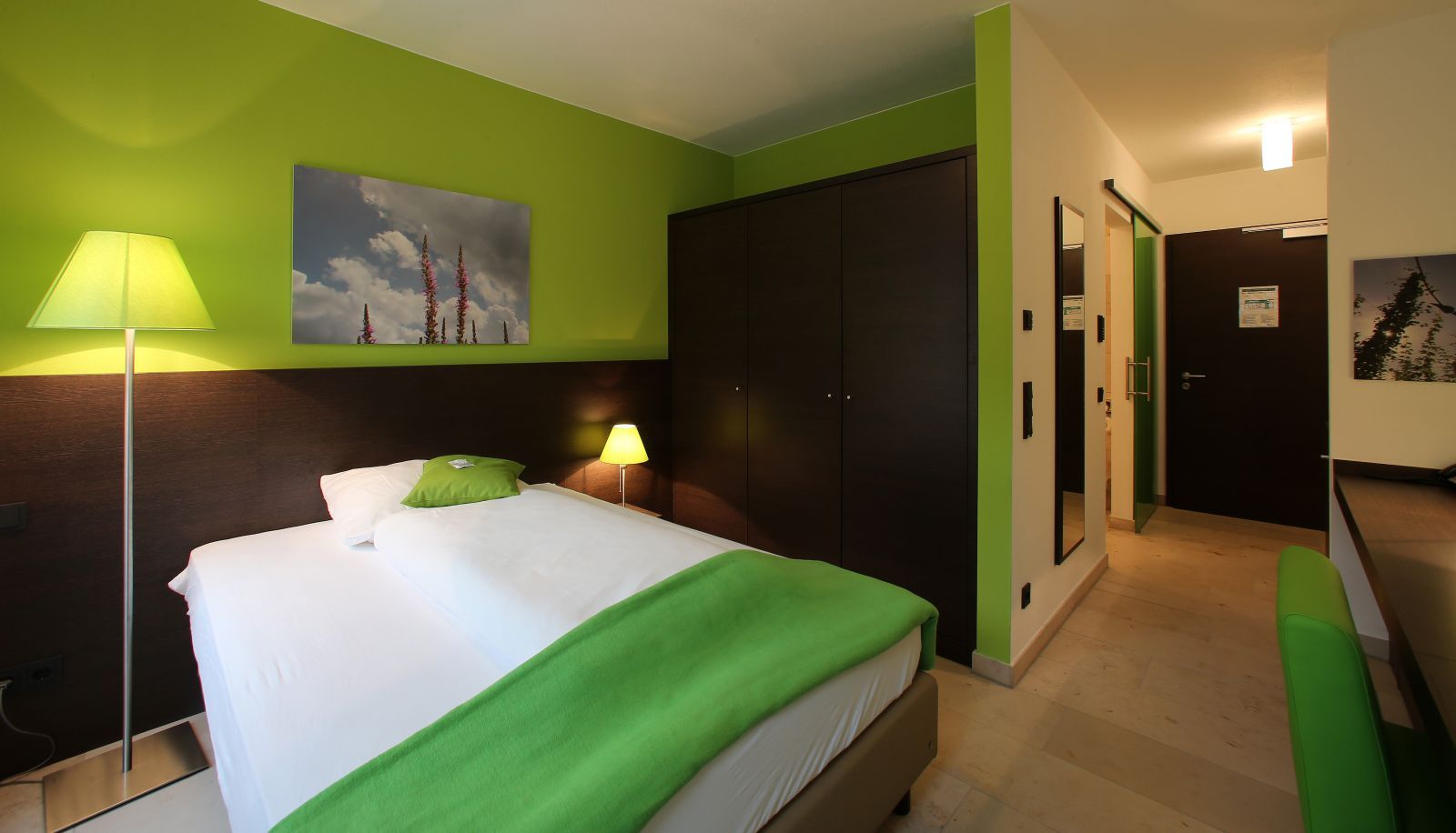 A hotel room with a bed, bedside lamps, a painting, a wardrobe, and a green chair.