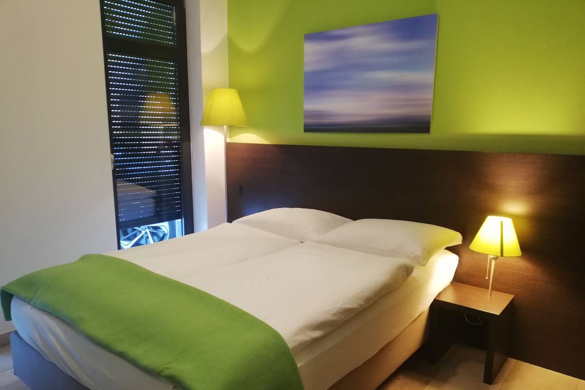 A hotel room with a bed, two bedside lamps, a green blanket, a painting on the wall, and a window with blinds.