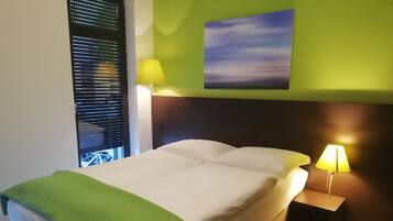 Habitación de hotel con cama, dos lámparas de noche, una manta verde, un cuadro en la pared y una ventana con persianas.