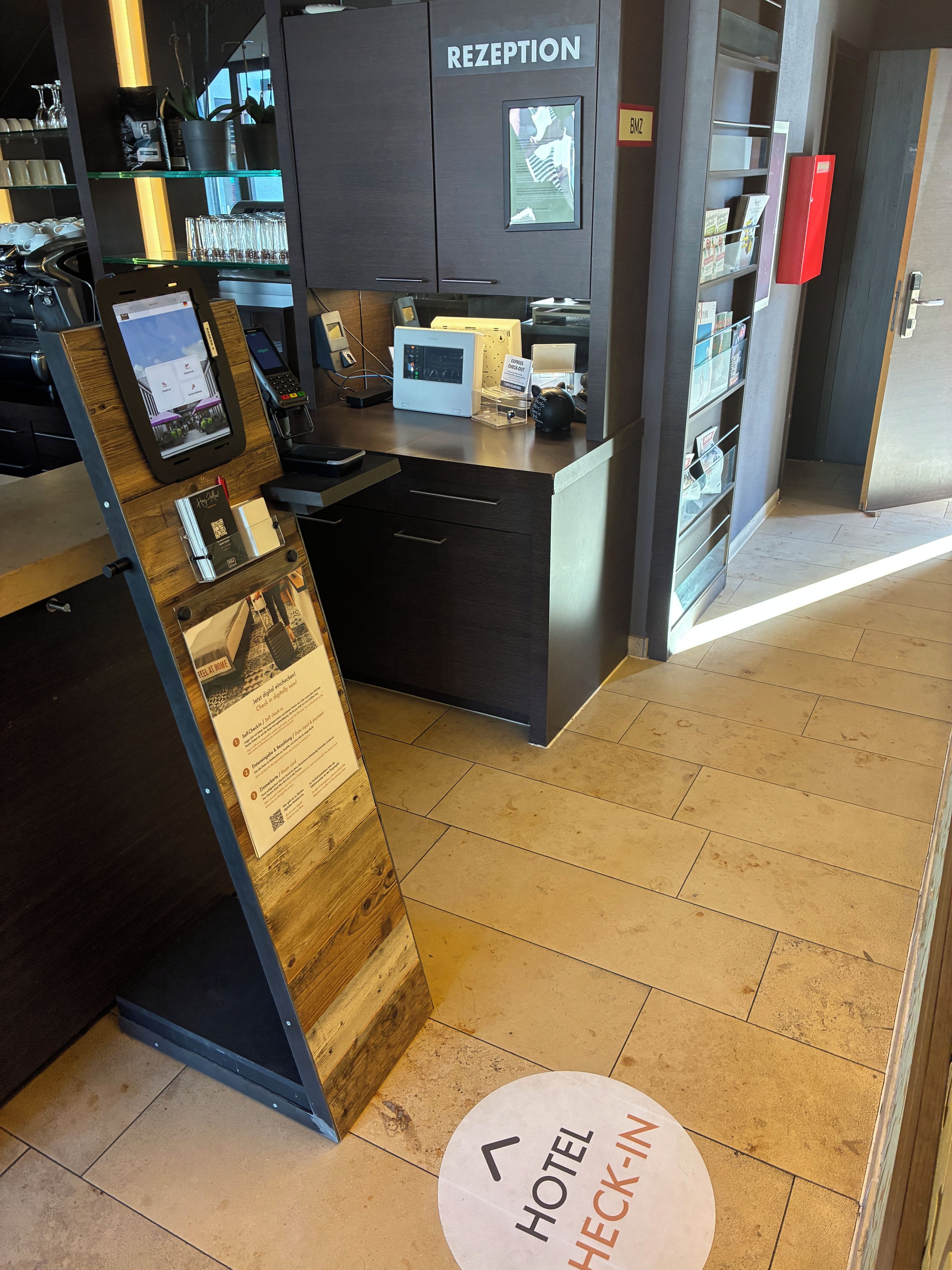 Un área de recepción con un quiosco digital, una mesa de recepción y un cartel que indica 'Hotel Check-in'.