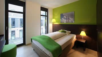 Una habitación de hotel con una cama, una silla verde, una mesita de noche con una lámpara y un cuadro en la pared.