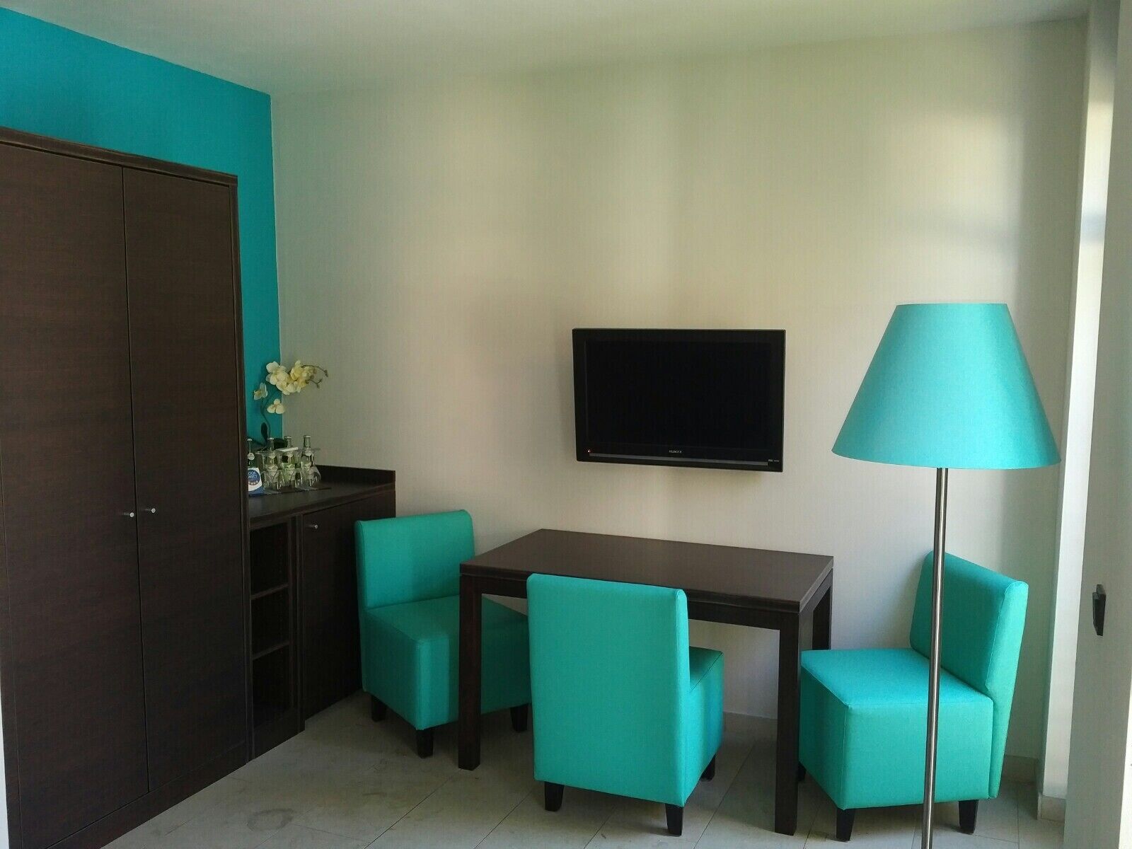 A small, modern room with teal walls, a dark wooden wardrobe, a flat-screen TV mounted on the wall, a teal floor lamp, and a table with teal chairs.