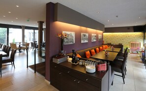 A modern dining area with a long table, cushioned seating, and a decorative wall with framed pictures. - KH Hotel (Geisenfeld)