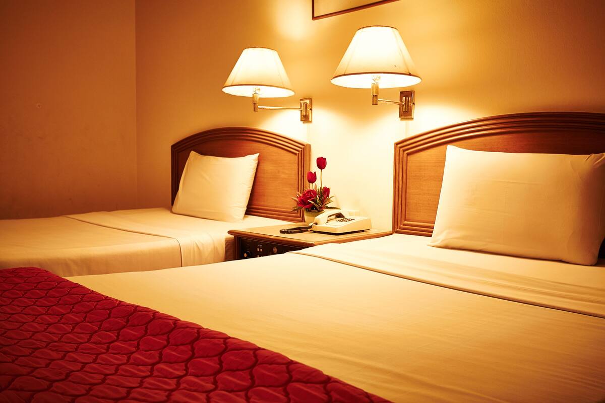 Habitación de hotel con dos camas, cada una con cabecero de madera, una mesita de noche con lámpara y un jarrón con flores rojas.
