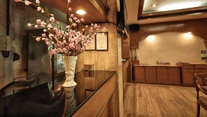 A reception area with a wooden reception desk, a vase with pink flowers, a wall-mounted menu, and a wooden cabinet in the background.