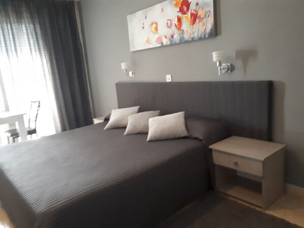 A modern hotel room with a large bed, two bedside tables, a painting on the wall, and a window with curtains. - Irene's Apartments (Limassol)