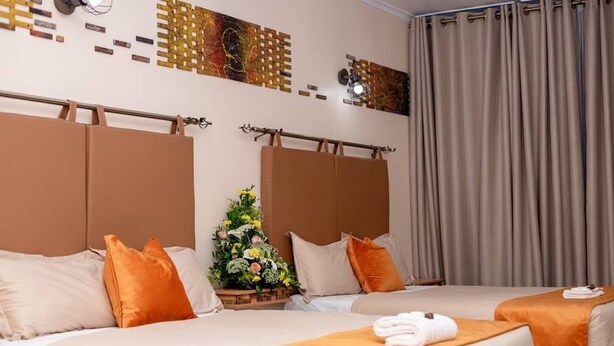 A hotel room with two beds, beige bedding, orange pillows, a small floral arrangement, and a curtain. - Bulawayo Central Lodge (Bulawayo)