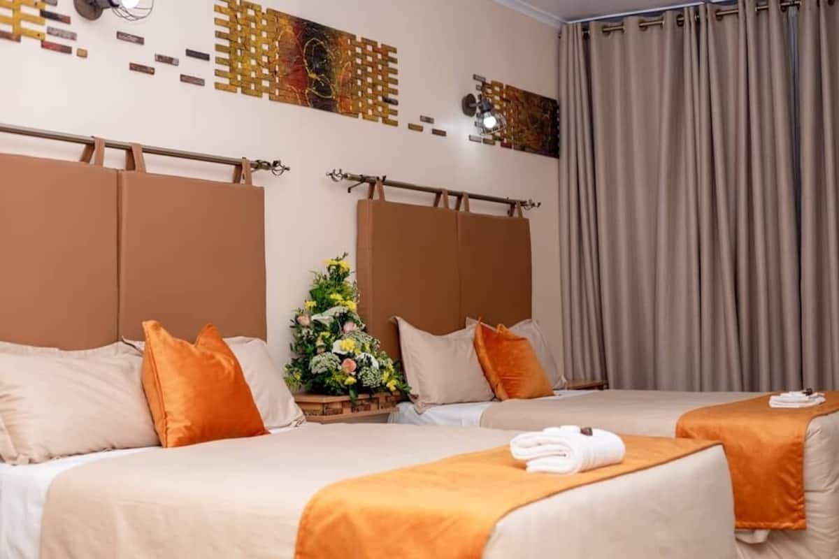 A hotel room with two beds, beige bedding, orange pillows, a small floral arrangement, and a curtain.