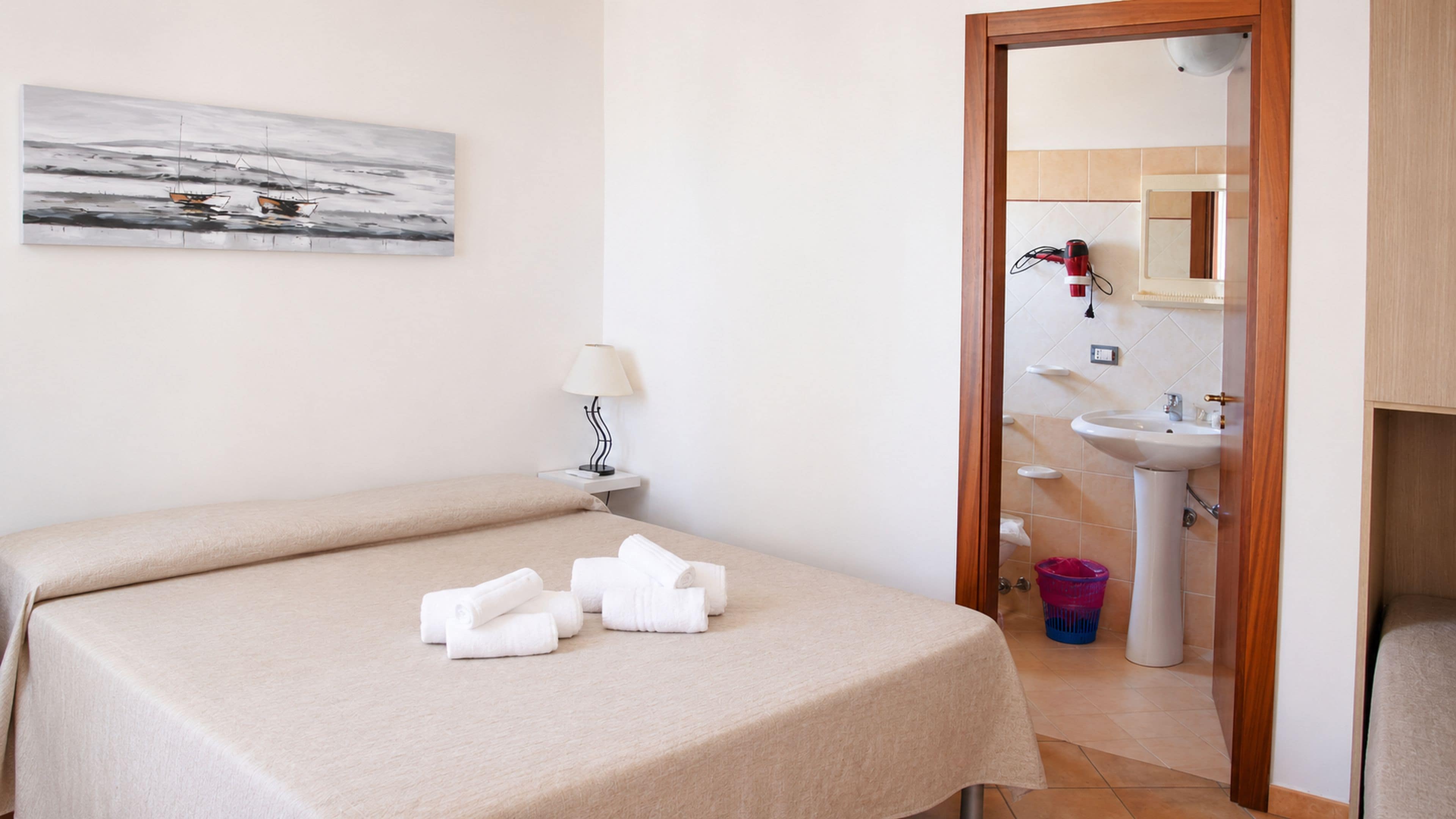 Une chambre d’hôtel avec un lit, une table de chevet avec une lampe, une salle de bain visible à travers une porte ouverte et un tableau accroché au mur.