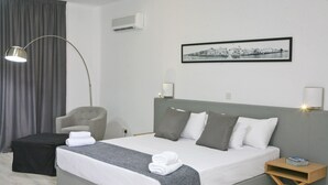 Una habitación de hotel moderna con una cama grande, ropa de cama gris y blanca, un cabecero gris, una mesita de noche con una lámpara, una silla, un reposapiés negro y un cuadro con una vista urbana en la pared.