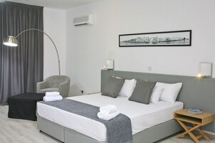 A modern hotel room with a large bed, grey and white bedding, a grey headboard, a side table with a lamp, a chair, a black ottoman, and a framed cityscape picture on the wall.