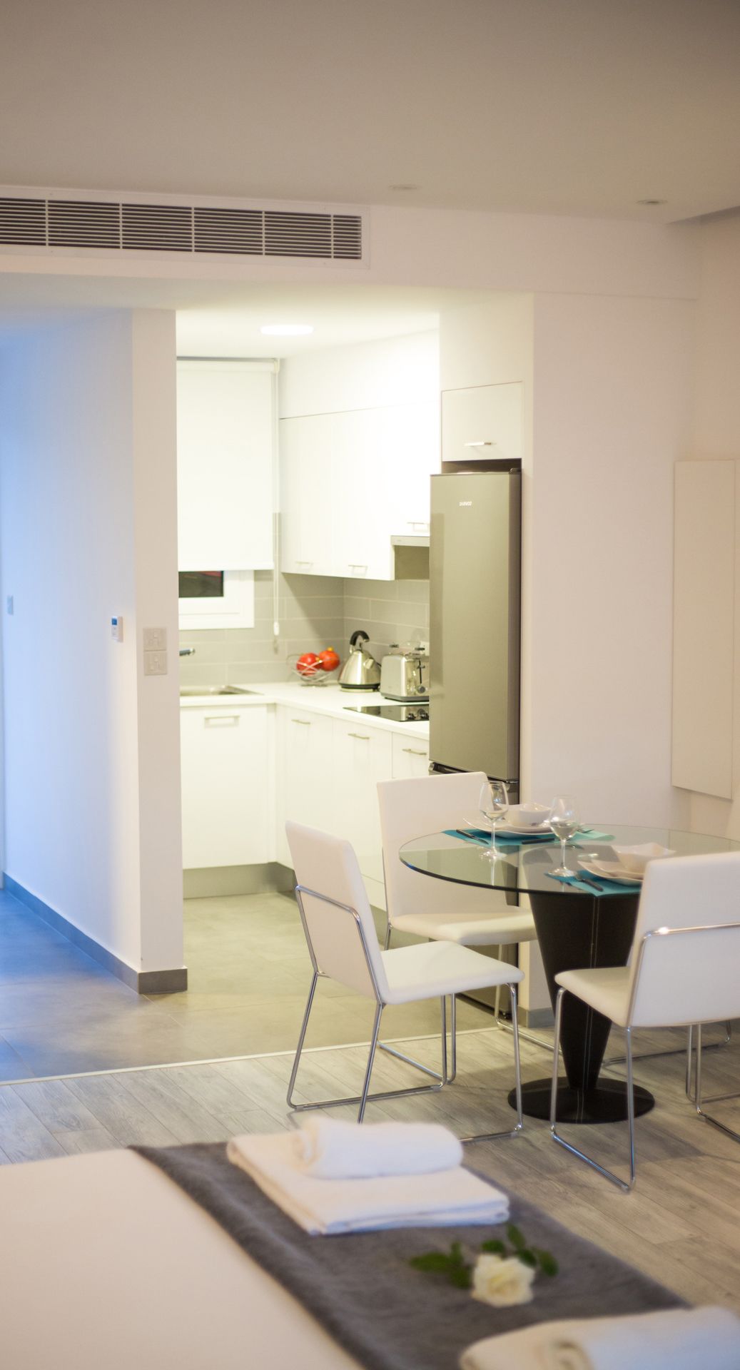 A modern hotel room with a dining area featuring a glass table and white chairs, a kitchen with white cabinets and a stainless steel refrigerator, and a visible air vent on the ceiling.