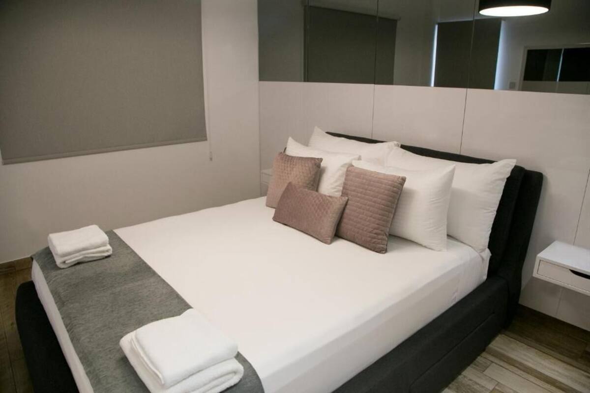 A neatly made bed with white and brown pillows, a grey blanket, and folded towels on a grey platform.