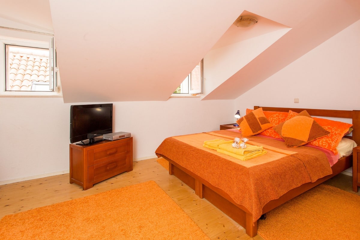 A bedroom with a sloped ceiling, a bed with orange bedding, a TV on a wooden dresser, and a window with a curtain.