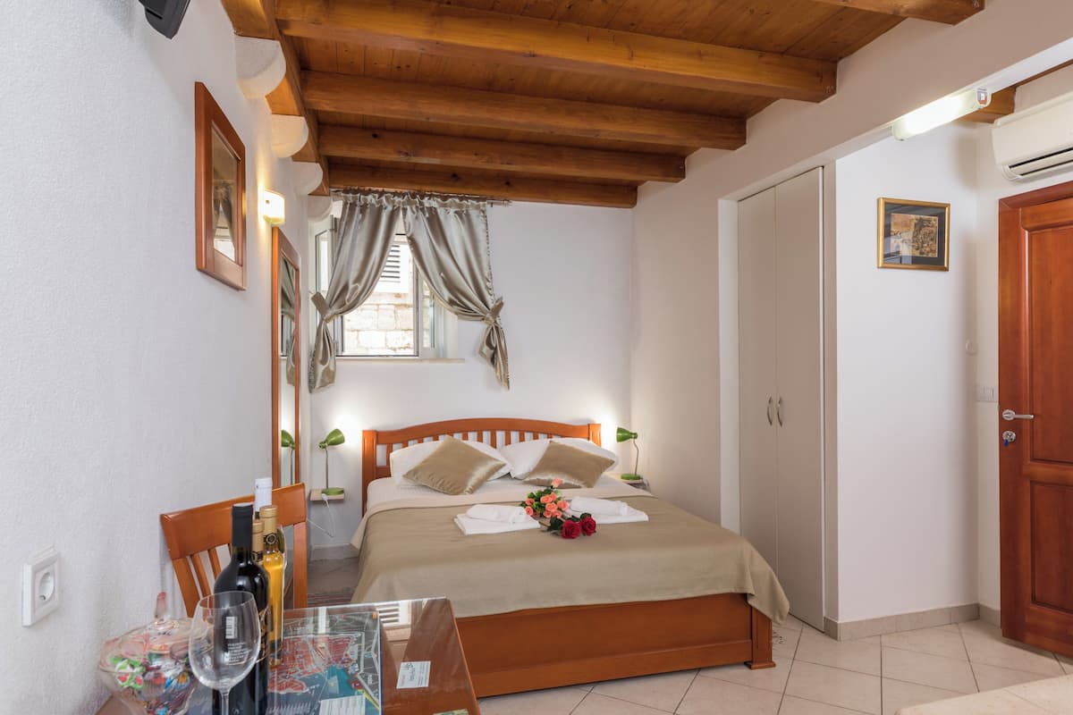 A bedroom with a wooden bed, a bedside table with a bottle and glass, a window with curtains, a wardrobe, and a door.