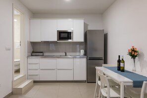 Cocina compacta con armarios blancos, microondas, refrigerador, lavaplatos y una mesa de comedor con sillas. Hay dos botellas de vino sobre la mesa.
