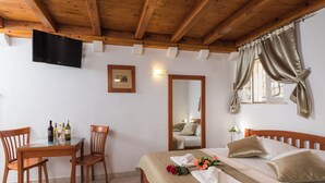A hotel room with a wooden ceiling, a bed with beige bedding, a wooden desk with chairs, a flat-screen TV mounted on the wall, and a window with curtains. - Apartments Ivana (Dubrovnik)