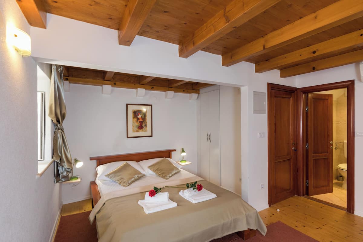 A bedroom with a wooden ceiling, a bed with beige bedding, a bedside lamp, a framed picture on the wall, and a wooden door leading to a bathroom.