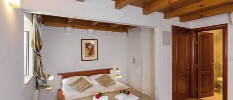 Un dormitorio con techo de madera, una cama con ropa de cama color beige, una lámpara de noche, un cuadro enmarcado en la pared y una puerta de madera que da a un baño.