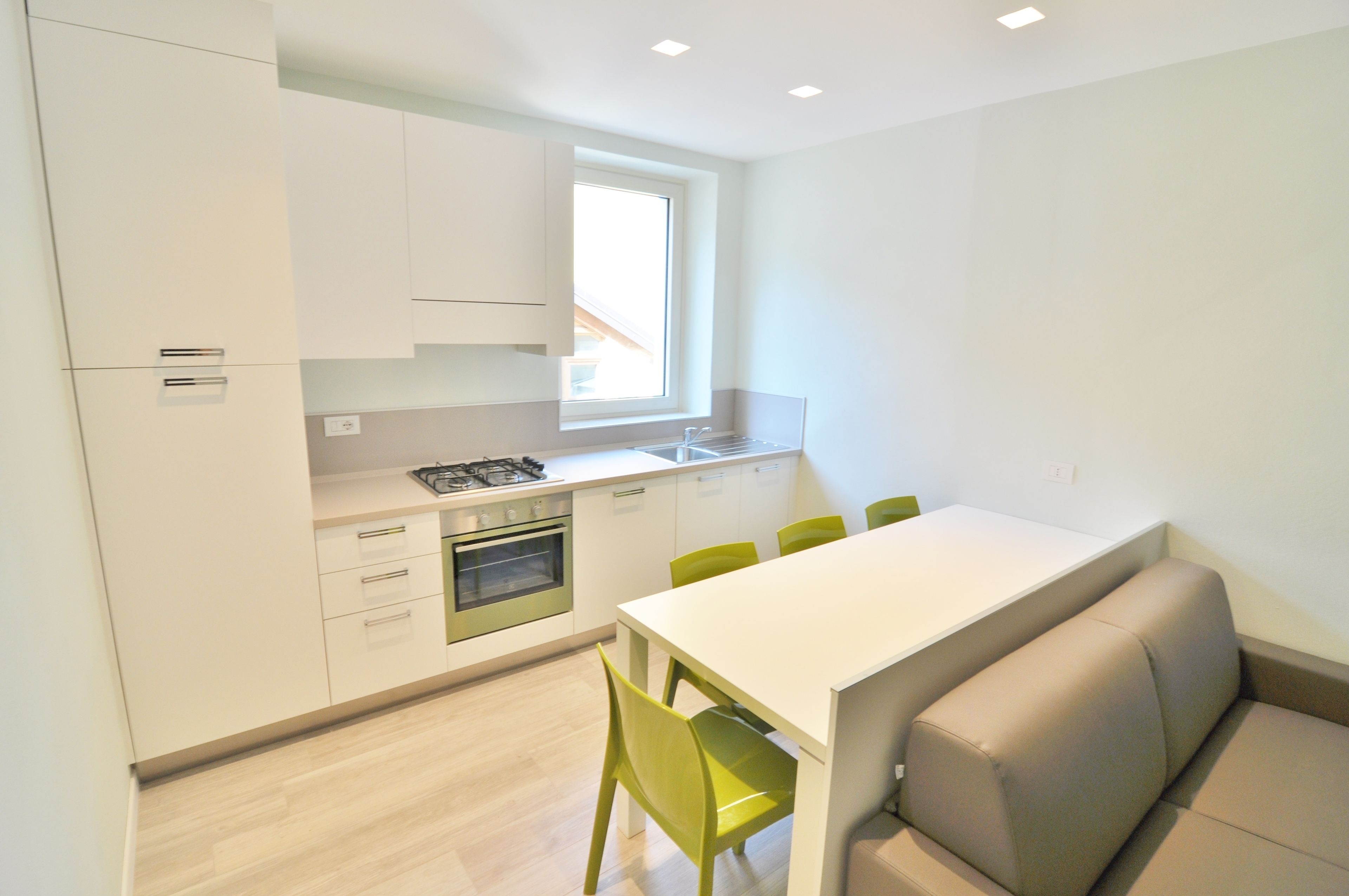 Una cocina moderna con armarios blancos, horno empotrado y ventanal sobre el fregadero. Hay una mesa de comedor con sillas verdes y un banco.
