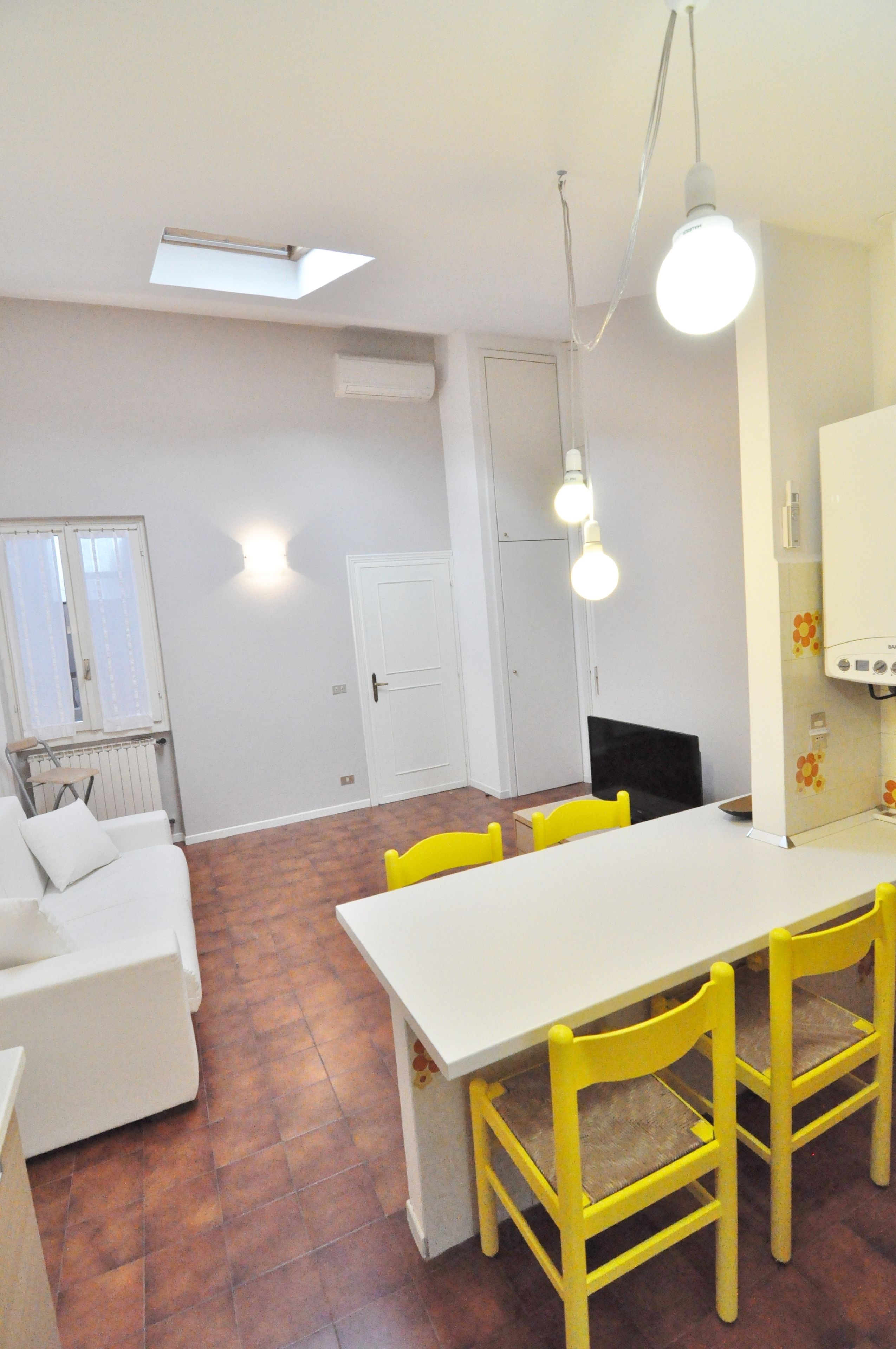 A room with a white sofa, a dining table with yellow chairs, and a white radiator.