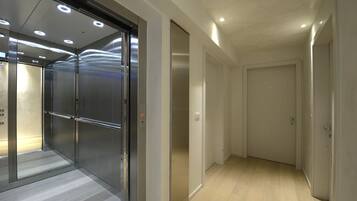 Un ascensor moderno con puertas de cristal y estructura metálica, un pasillo con suelo de madera y una puerta blanca sencilla al final.