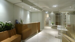 Una moderna zona de recepción con un panel de vidrio, mostrador de madera y un pequeño rincón de descanso con plantas.