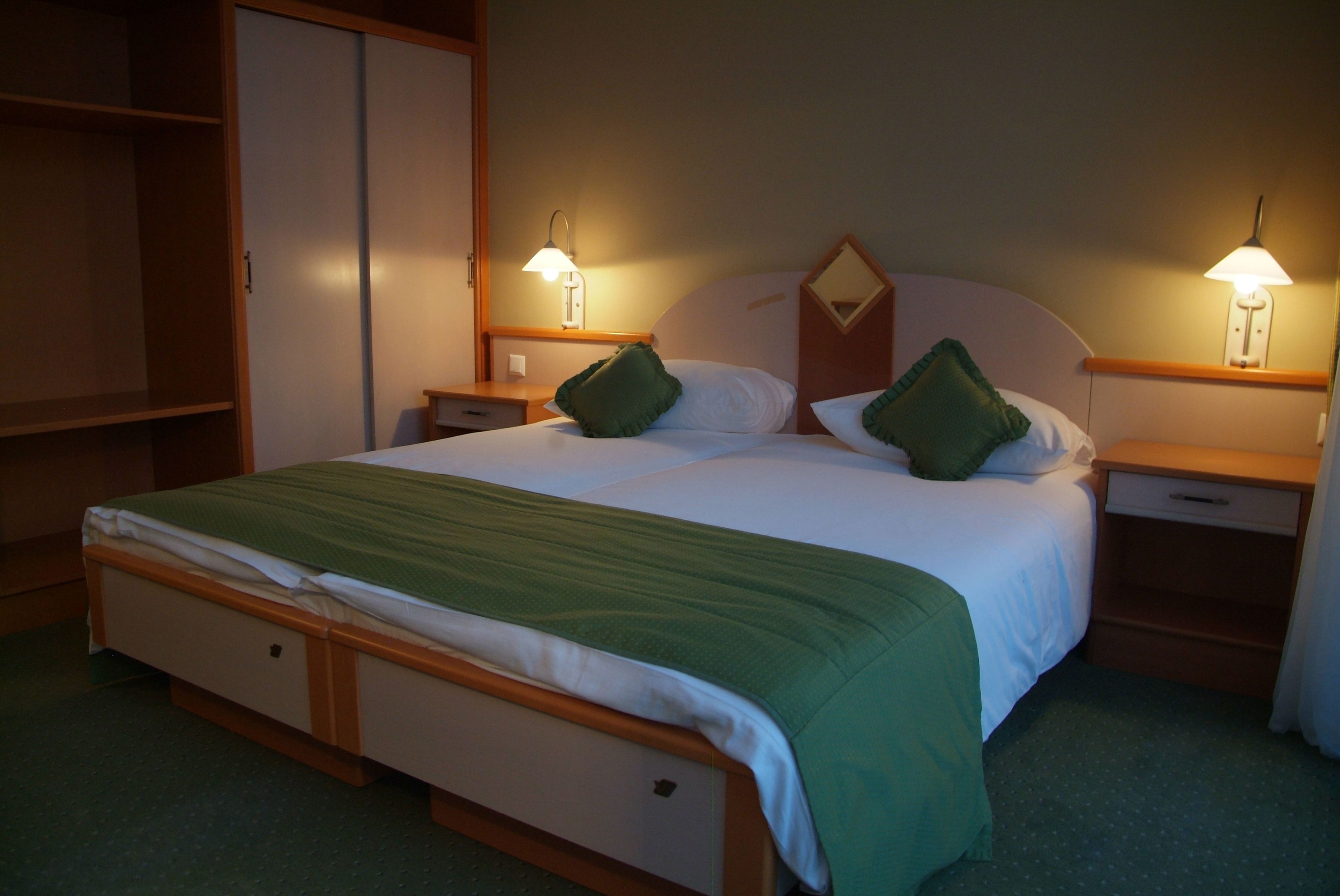 A hotel room with a single bed, two bedside tables, a nightstand, a wardrobe, and two wall-mounted lamps.