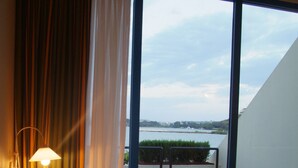 Quarto de hotel com cama, mesa de cabeceira, luminária e vista para um corpo de água através de grandes janelas.