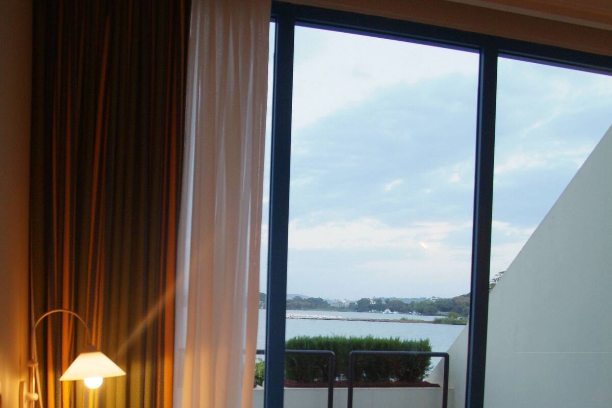 Ein Hotelzimmer mit einem Bett, einem Nachttisch, einer Lampe und Blick auf ein Gewässer durch große Fenster.