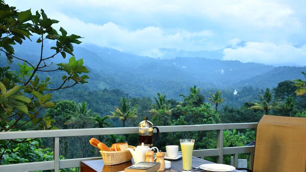 A table with breakfast items, a book, and a view of a lush landscape. - Mistletoe Munnar (Devikolam)