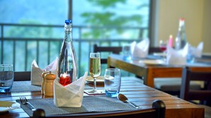 A dining table set with a bottle, glasses, and cutlery, with a view of greenery through a window. - Mistletoe Munnar (Devikolam)