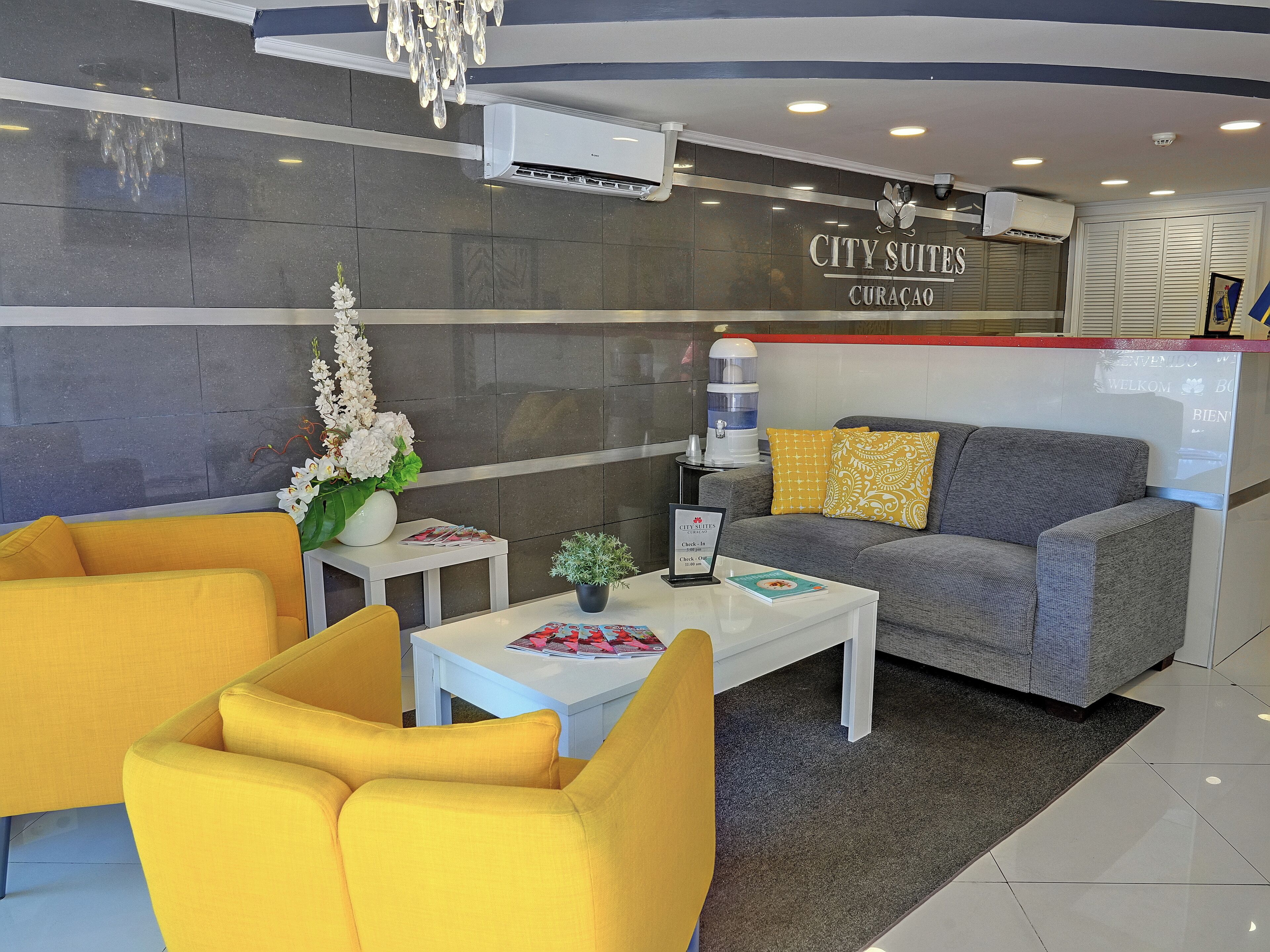 Un moderno vestíbulo de hotel con un sofá gris, sillones amarillos, una mesa de centro blanca y un mostrador de recepción.