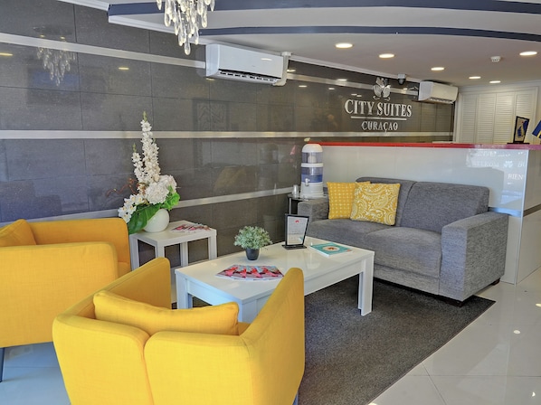 Un hall d’hôtel moderne avec un canapé gris, des fauteuils jaunes, une table basse blanche et un comptoir d’accueil.