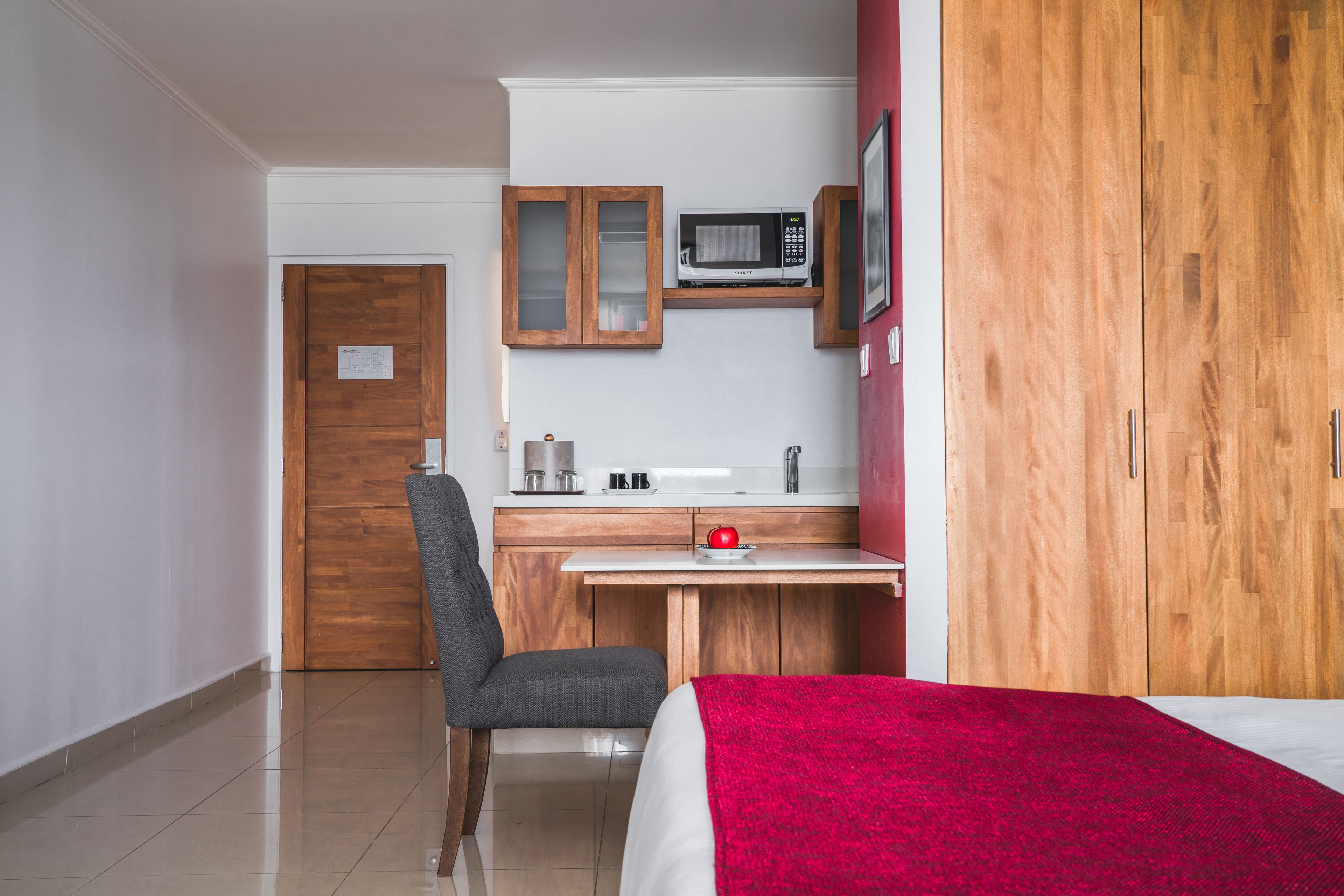 A compact hotel room with a wooden wardrobe, a kitchenette with a microwave, a small desk, and a chair.