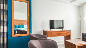 A modern hotel room with a blue accent wall, a wooden desk with a flat-screen TV, a large mirror, and two upholstered chairs.