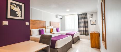 A hotel room with two beds, a dining table, and a chair. There is a framed picture on the wall and a window with curtains.
