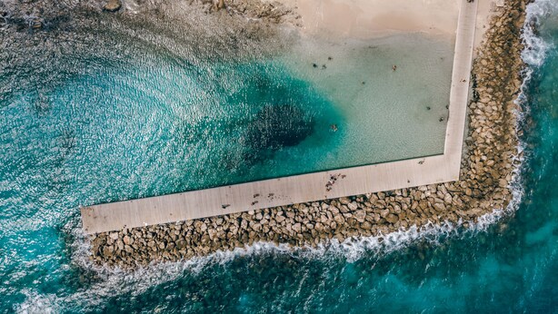 An aerial view of a beach with clear turquoise waters, a rocky shoreline, and a wooden walkway leading to the water. - Majestic City Suites & Beach Hotel (Willemstad)